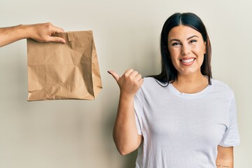 Sticker - Beautiful brunette woman with take away paper bag pointing thumb up to the side smiling happy with open mouth