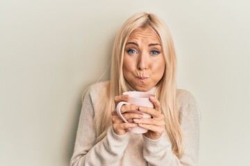 Canvas Print - Young blonde woman drinking a cup of coffee puffing cheeks with funny face. mouth inflated with air, catching air.