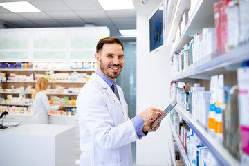 Wall Mural - Pharmacist standing by the shelves with medicines and typing on tablet in drug store.