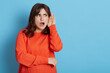 © sementsova321 - Shocked female wearing orange casual sweater posing with palm near ear, trying to hear secret information, gossip, woman with astonished expression, isolated over blue background.