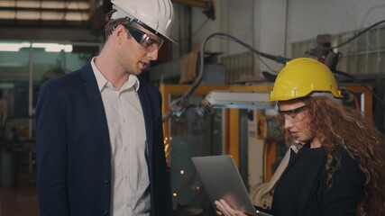 Two heavy industry business engineers in hardhats discuss information on laptop computer while standing indoors welding manufacturing industrial