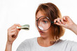 Portrait of a woman looks through a magnifying glass at a small wad of dollars. White background. The concept of checking counterfeit money