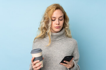 Poster - Young blonde woman wearing a sweater isolated on blue background holding coffee to take away and a mobile