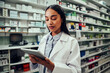 © StratfordProductions - Young female pharmacist working using digital tablet standing in chemist wearing labcoat