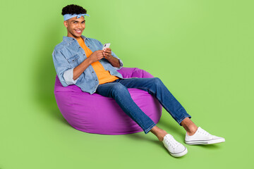 Sticker - Full length body size photo of young guy in beanbag browsing internet on smartphone social media isolated on bright green color background