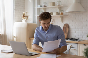 Wall Mural - Focused man working with documents, paying bills, sitting at table with laptop in modern kitchen, businessman freelancer reading correspondence, checking, analyzing financial report, paperwork