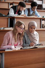 Poster - Two intercultural female students of college or university sitting on the first row by desk in lecture hall and discussing passage in book at lesson