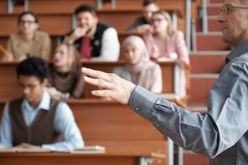 Wall Mural - Hand of mature professor in casualwear standing in front of audience and explaining subject of lecture to young intercultural students