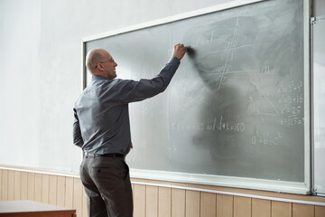 Poster - Bald professor of university in smart casualwear standing in front of blackboard and writing down algebraic formula during explanation