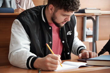 Poster - Serious guy in casualwear bending over desk and making notes in copybook while scrolling in smartphone during lecture in university