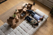 © fizkes - Top view happy couple tenants homeowners relaxing on couch on moving day in living room with cardboard boxes, man and woman resting after relocation in first own apartment, mortgage or rent