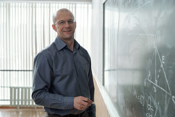 Poster - Successful mature teacher or professor in smart casualwear holding piece of chalk while standing by blackboard in front of camera in classroom