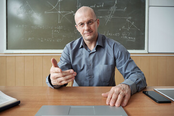 Poster - Mature teacher in shirt and eyeglasses sitting by desk, looking at laptop display in front of him and explaining subject at lesson in classroom