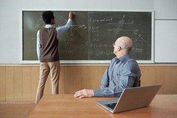Poster - Mature bald professor sitting by desk and looking at student standing by blackboard, drawing graph and explaining his actions at lesson