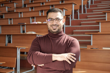 Poster - Young successful male teacher of university crossing his arms by chest while standing in front of camera against desks in lecture hall