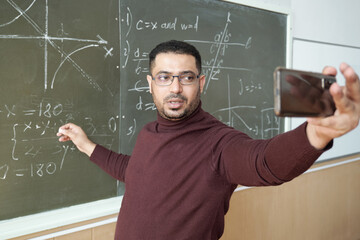 Poster - Mixed-race male in casualwear holding smartphone in front of himself while standing by blackboard and explaining how to solve equation