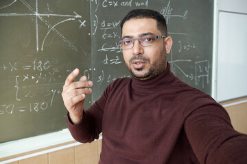 Aufkleber - Mixed-race man holding smartphone in front of himself and looking at you while standing by blackboard and explaining how to solve equation
