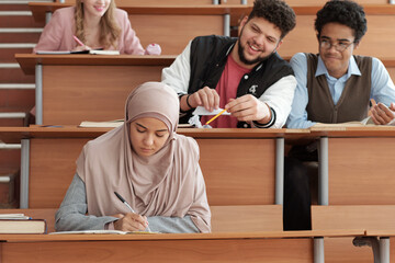 Poster - Serious girl in hijab making notes in copybook at lecture while sitting by desk against two intercultural groupmates making fun of her
