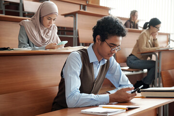 Poster - Mixed-race guy and other college students in casualwear sitting by wooden desks in lecture hall at lesson and scrolling in smartphones