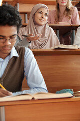 Poster - Young pretty female student in hijab sitting by desk during lecture and looking at teacher while listening to him among her groupmates