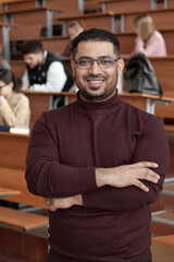 Poster - Young successful male teacher crossing his arms by chest while standing in front of camera against students sitting by desks in lecture hall