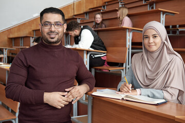 Poster - Successful male teacher standing by desk where young female student in hijab sitting and making notes while reading book at lesson