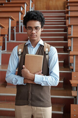 Poster - Pensive or offended intercultural male student in smart casualwear and eyeglasses holding book while standing against staircase in auditorium