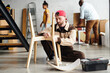 Contemporary furniture assemblage specialist in workwear sitting on the floor and fixing parts of wooden chair against family with boxes