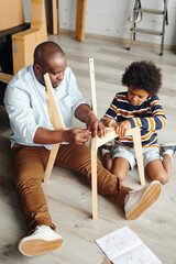 Canvas Print - Young man of African ethnicity teaching his cute little son how to assemble new furniture while sitting on the floor of new flat or house