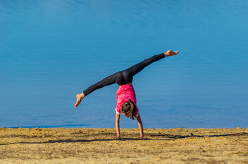 7 years old girl with pink shirt and sportswear doing a cartwheel at the beach during spring with blue water as background. Palencia, Spain