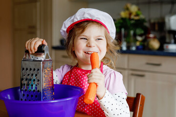 Cute little preschool girl grate carrots for baking carrot cake or cookies. Happy toddler child in apron and chef hat help in the kitchen prepare healthy salad lunch, indoors. Healthy food with kids.