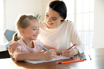 Loving young Caucasian mother and little 7s daughter have fun drawing together in album a home. Happy caring mom and small girl child engaged in funny creative activity together. Art, hobby concept.