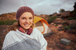 © StratfordProductions - Smiling woman with blanket on mountain standing outside tent during camping at mountain hill