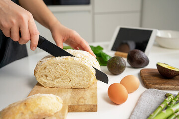 Woman hand holding black knife and cutting bread for breakfast toasts. Eggs, avocado, asparagus and tablet with recipe on white table in home kitchen.