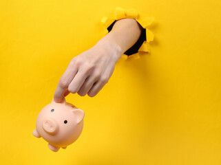 Poster - Female hand holds piggy bank through torn hole yellow paper. Concept art. Minimalism