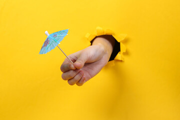 Poster - Female hand holds cocktail umbrella through torn hole yellow paper. Concept art. Minimalism.