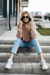 Poster - Fasion blonde woman in brown oversize hoodie, glasses and blue jeans posing sitting on the steps.  Mockup for logo or branding design