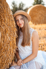 Canvas Print - Beautiful young girl in a white dress and a straw hat sitting near the bale of straw. Outdoor portrait of a positive female