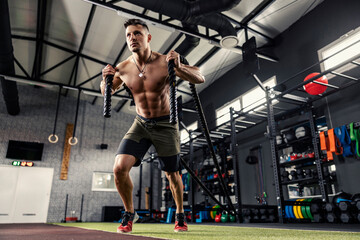 A muscular man without a shirt pulls a rope in a modern gym with equipment. A strong sportsman full of energy prepares to exercise in functional training. Getting ready for the fitness challenge