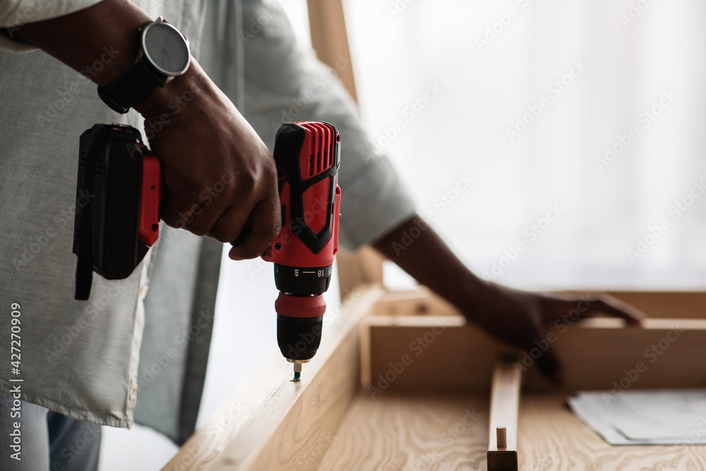 Unrecognizable black man assembling table, screwing details of desk with electric drill, closeup, crop - obrazy, fototapety, plakaty 