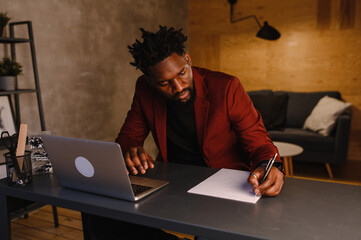 Handsome Black African American Man Working on Laptop Computer while Sitting Behind Desk in Cozy Living Room. Freelancer Working From Home