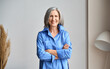 © insta_photos - Happy laughing mature elder 60s business woman standing indoors. Senior mid age older stylish look woman portrait with crossed arms looking at camera at modern home office.