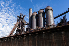 Rusting Abandoned Steel Mill, Blast Furnaces And Smokestacks Atop A Concrete Wall, Set Against A Brilliant Blue Sky, Horizontal Aspect