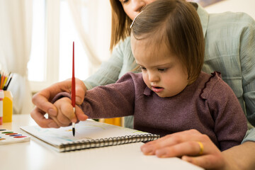 Kid with down syndrome drawing with paints on paper while sitting at the table