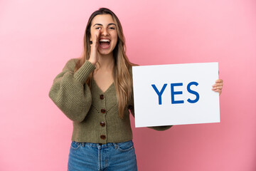Wall Mural - Young caucasian woman isolated on pink background holding a placard with text YES and shouting