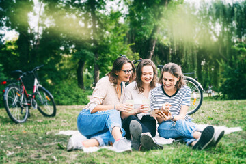 Wall Mural - Three young beautiful women friends having fun in a summer park sitting on green grass and looking into a mobile phone