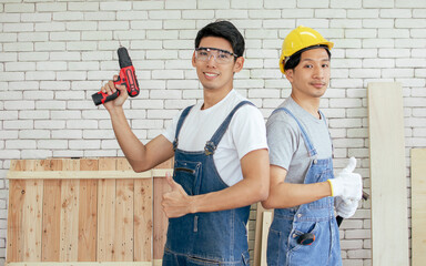 Portrait of two Asian handsome craftsmen wearing safety helmets, eyeglasses and blue overalls holding drill equipment and making DIY wooden furniture at home, posting and looking to camera.