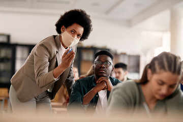 Wall Mural - African American professor wearing face mask while assisting her student on a class in the classroom.