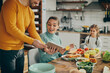 © Drazen - Happy woman watching tutorial on digital tablet while preparing meal for her family in dining room.