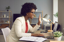Side View Of Friendly African American Business Woman Waving In Front Of Webcam Greeting Via Video Link. Female Employee Sits At A Desk In The Office And Communicates During An Online Conference.